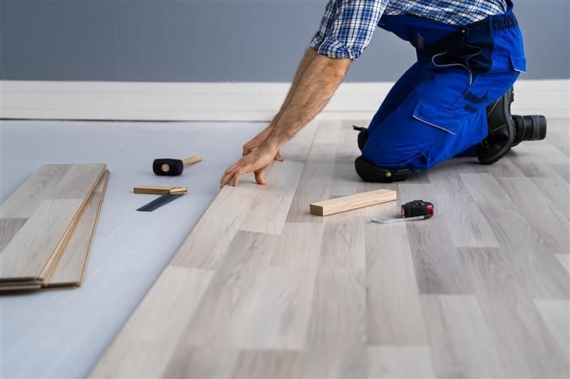 Man kneeling, installing light wood flooring. Tools and flooring are visible, indoors.