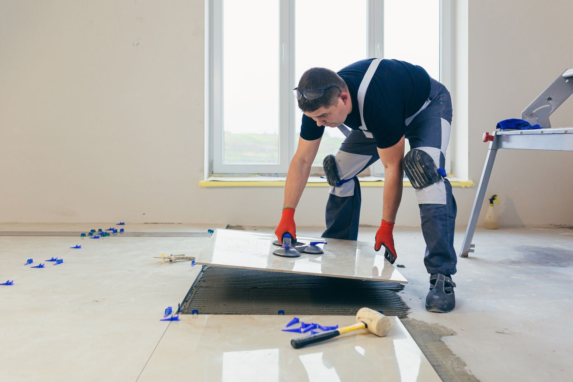 Man tiling a floor, using tools. Wearing knee pads and safety glasses.