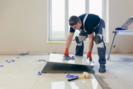 Man tiling a floor, using tools. Wearing knee pads and safety glasses.
