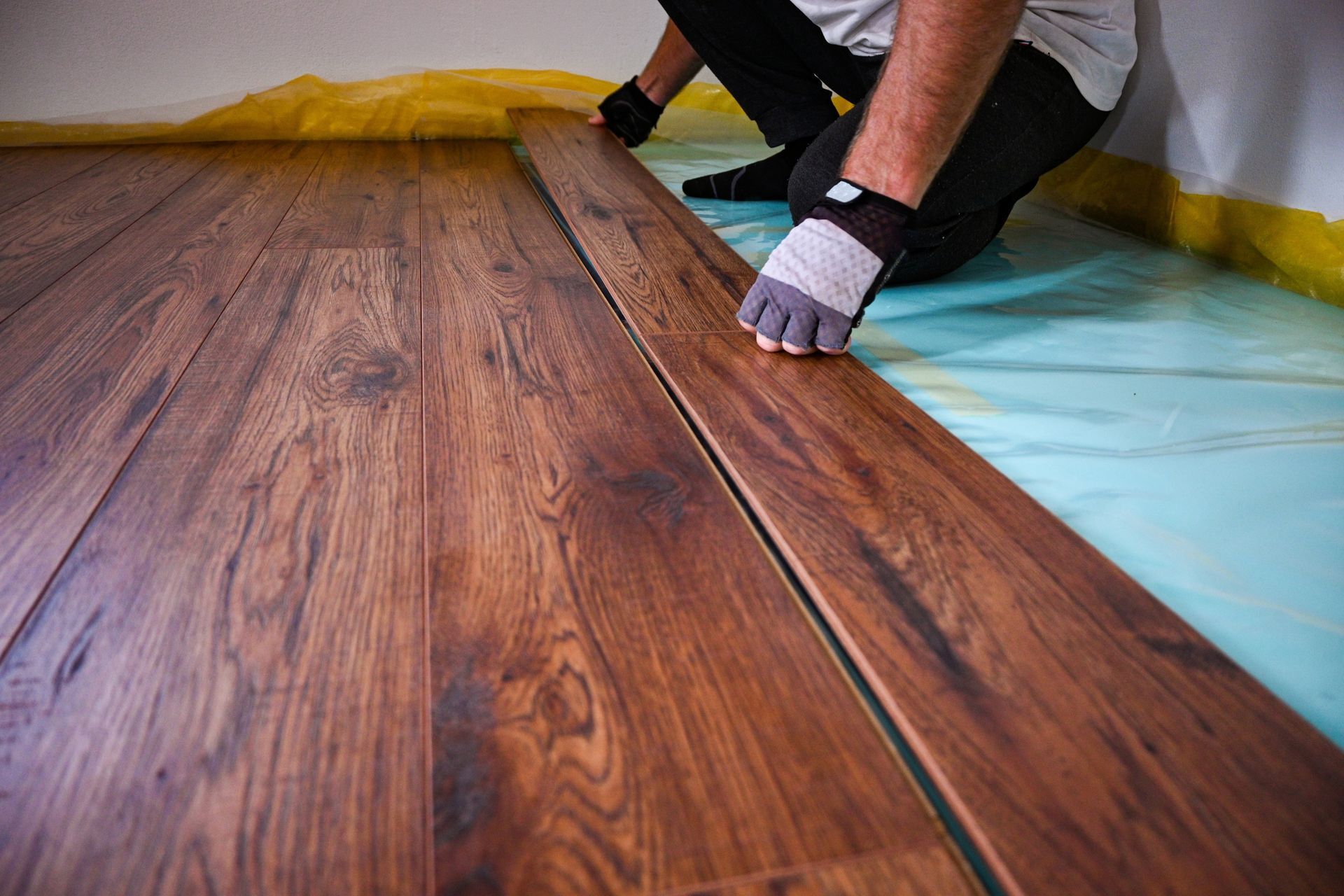 Person installing wood flooring, wearing gloves. Brown planks, blue and yellow underlayment.