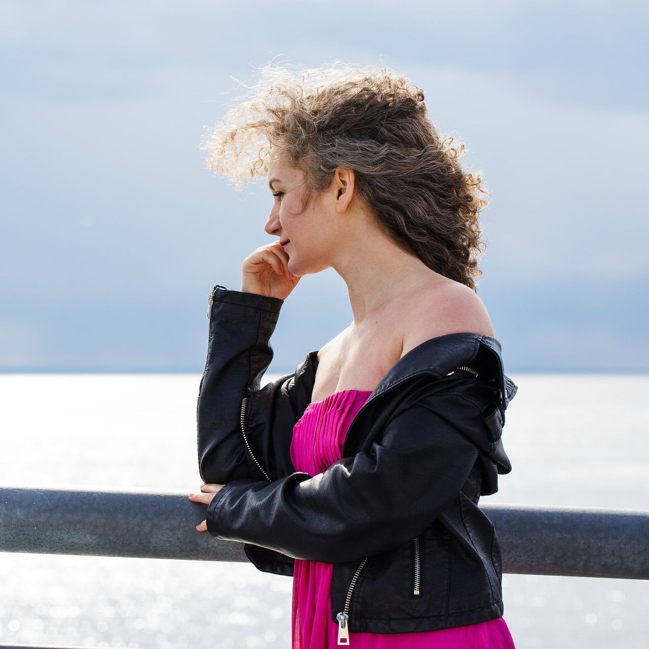 A woman in a pink dress and a black jacket stands on a pier overlooking the ocean