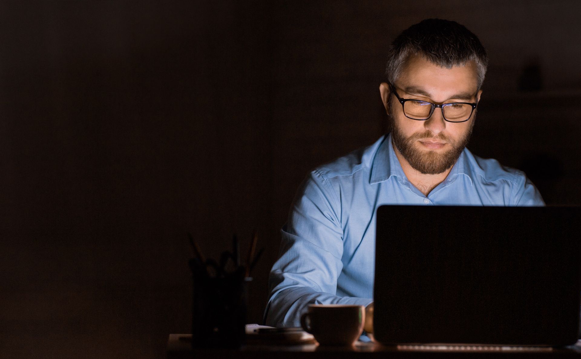 A man is sitting at a desk using a laptop computer in the dark.