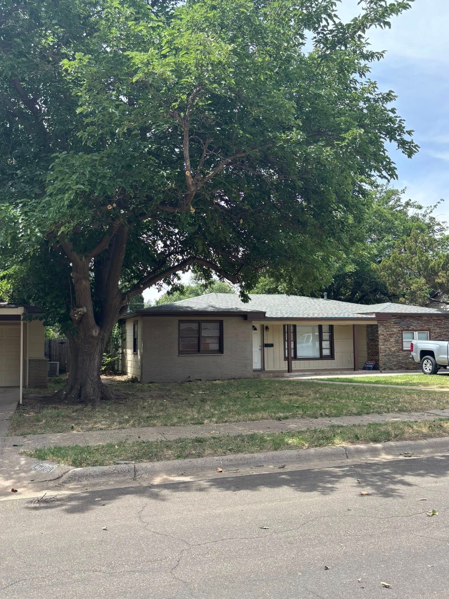A one-story beige house with a large tree in front; brown trim, porch, and a light-colored lawn.