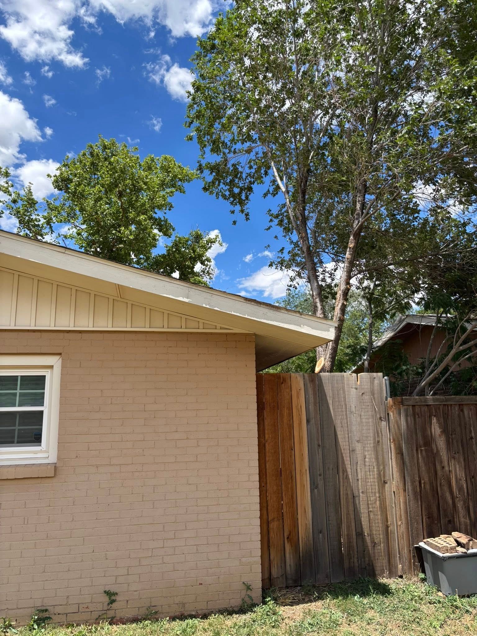 Tan brick building, wooden fence, and trees against a blue sky with fluffy clouds.