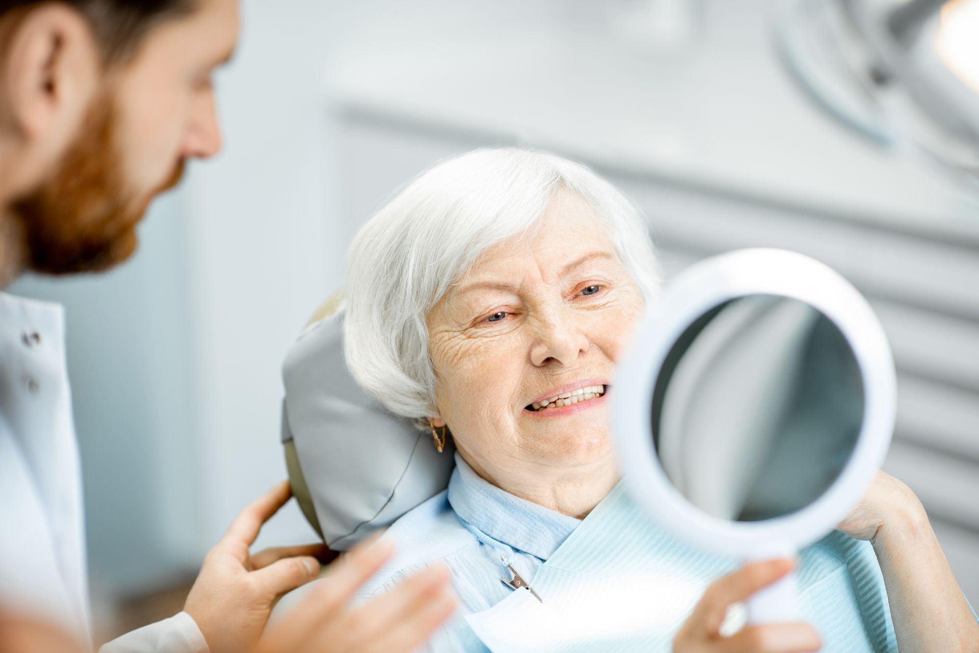 An elderly woman is sitting in a dental chair looking at her teeth in a mirror.