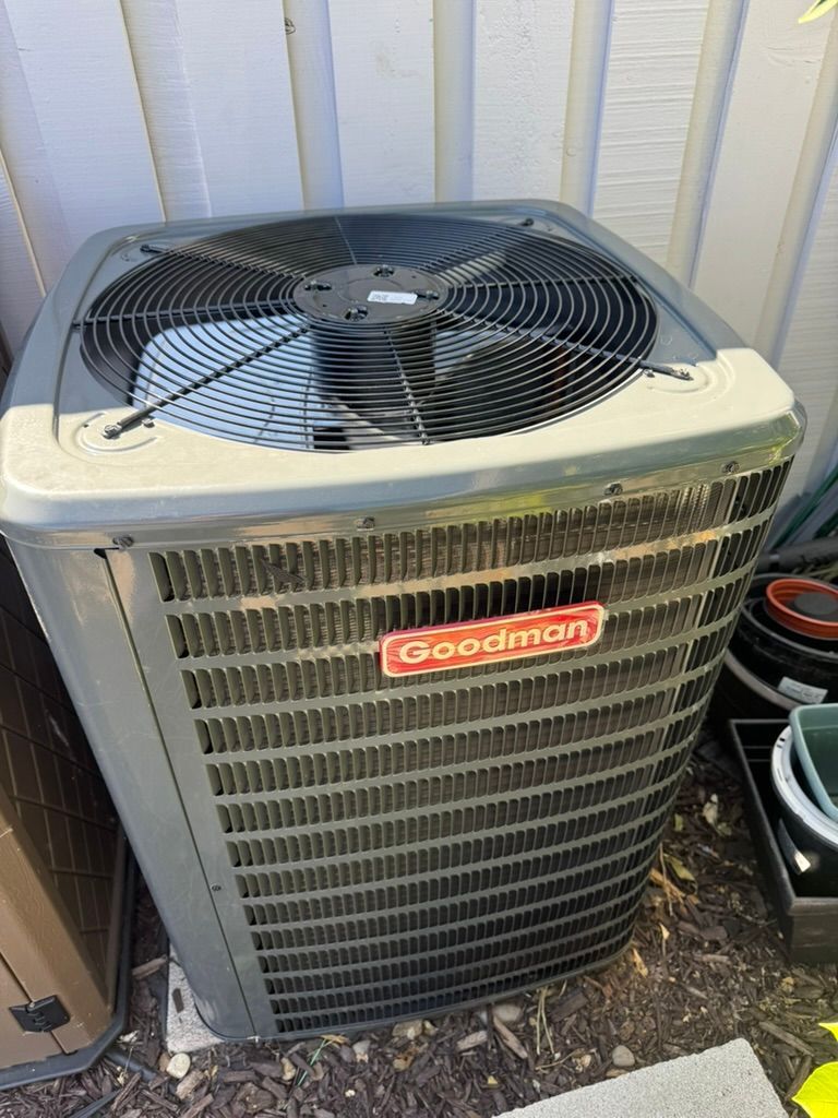 A goodman air conditioner is sitting on the ground in front of a white fence.