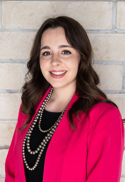 A woman wearing a pink jacket and a pearl necklace is smiling for the camera.
