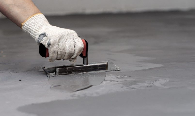 Hand wearing glove using trowel to smooth wet, gray cement floor.