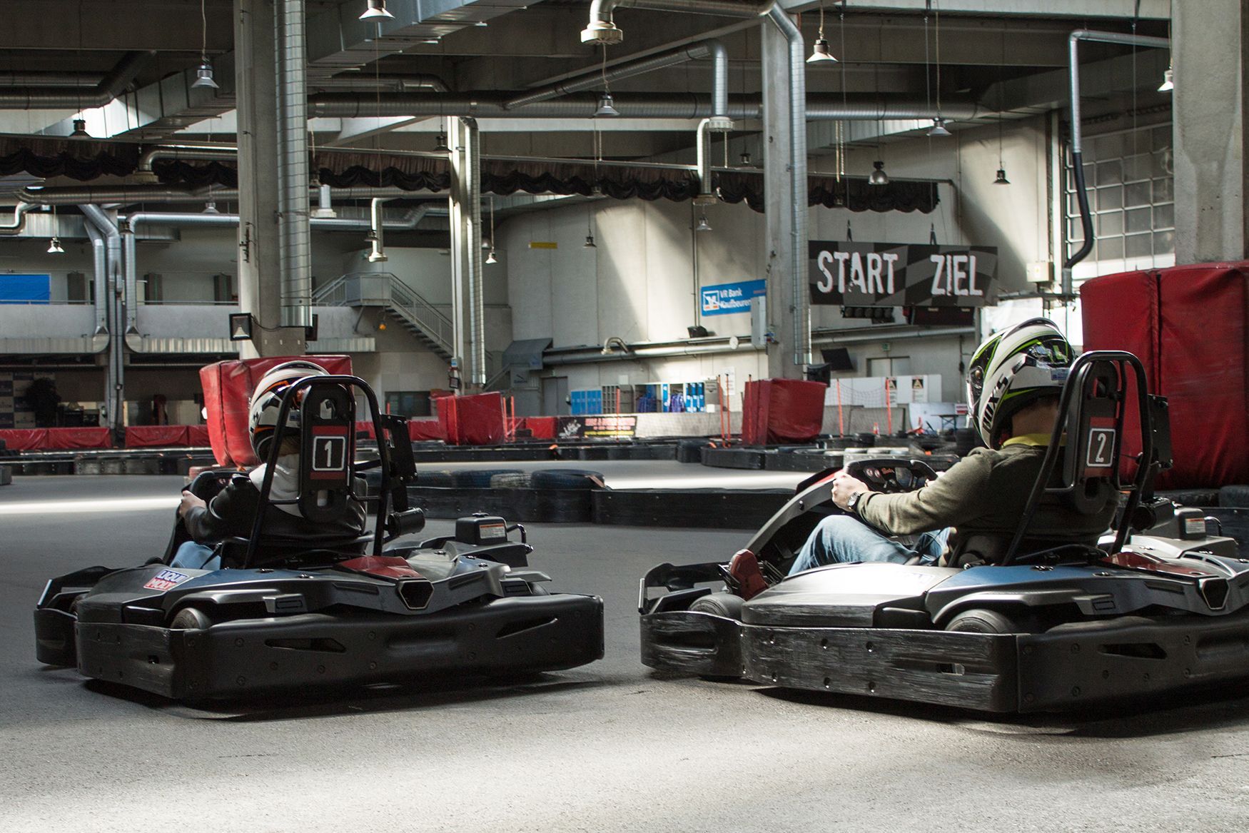 Zwei Personen fahren in schwarzen Go-Karts auf einer Indoor-Rennstrecke um die Wette; im Hintergrund ist ein Start-Tank-Schild zu sehen.