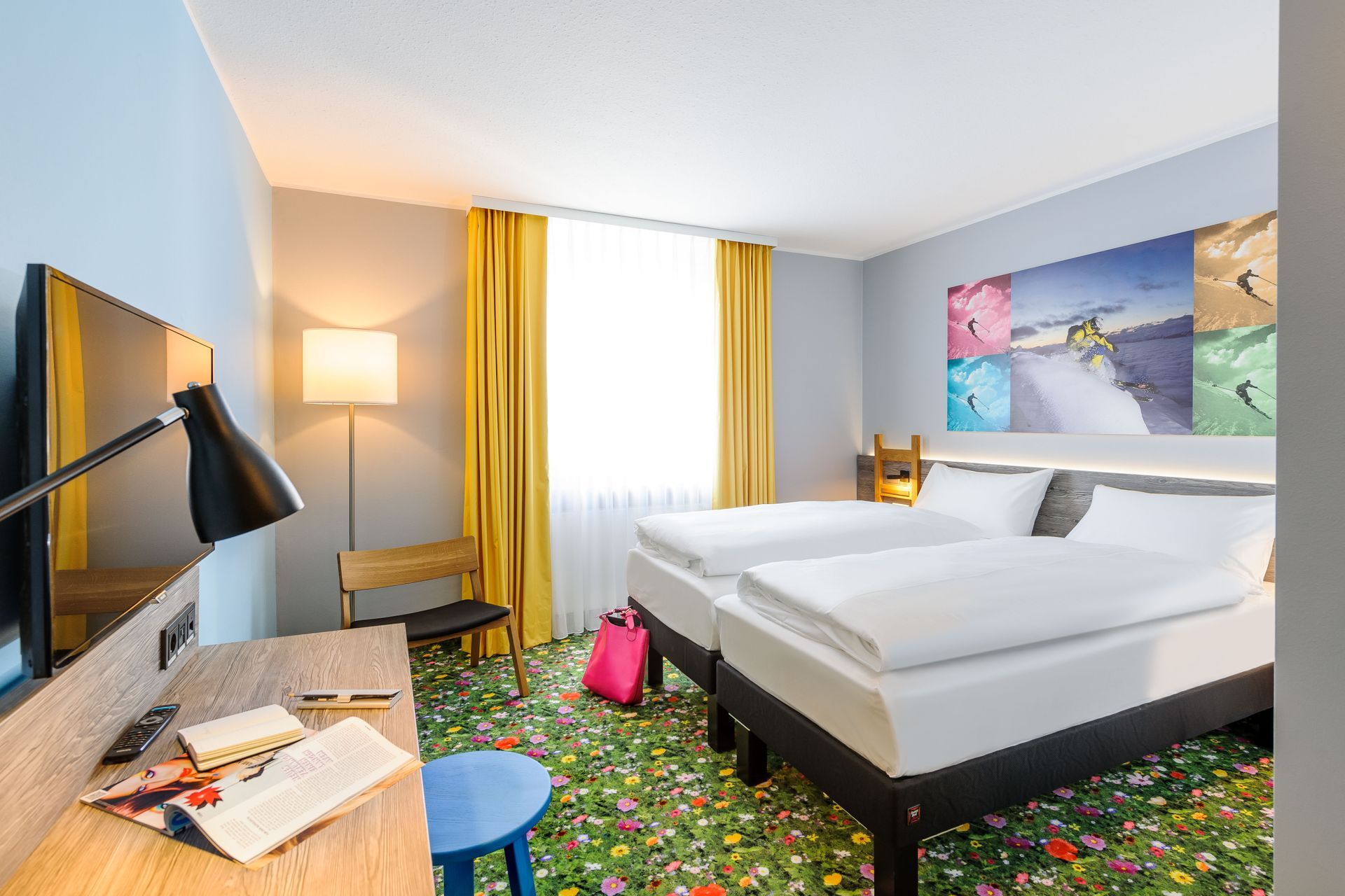 Modernes Hotelzimmer mit zwei weißen Betten, farbenfrohem Teppich, Schreibtisch und gelben Vorhängen.