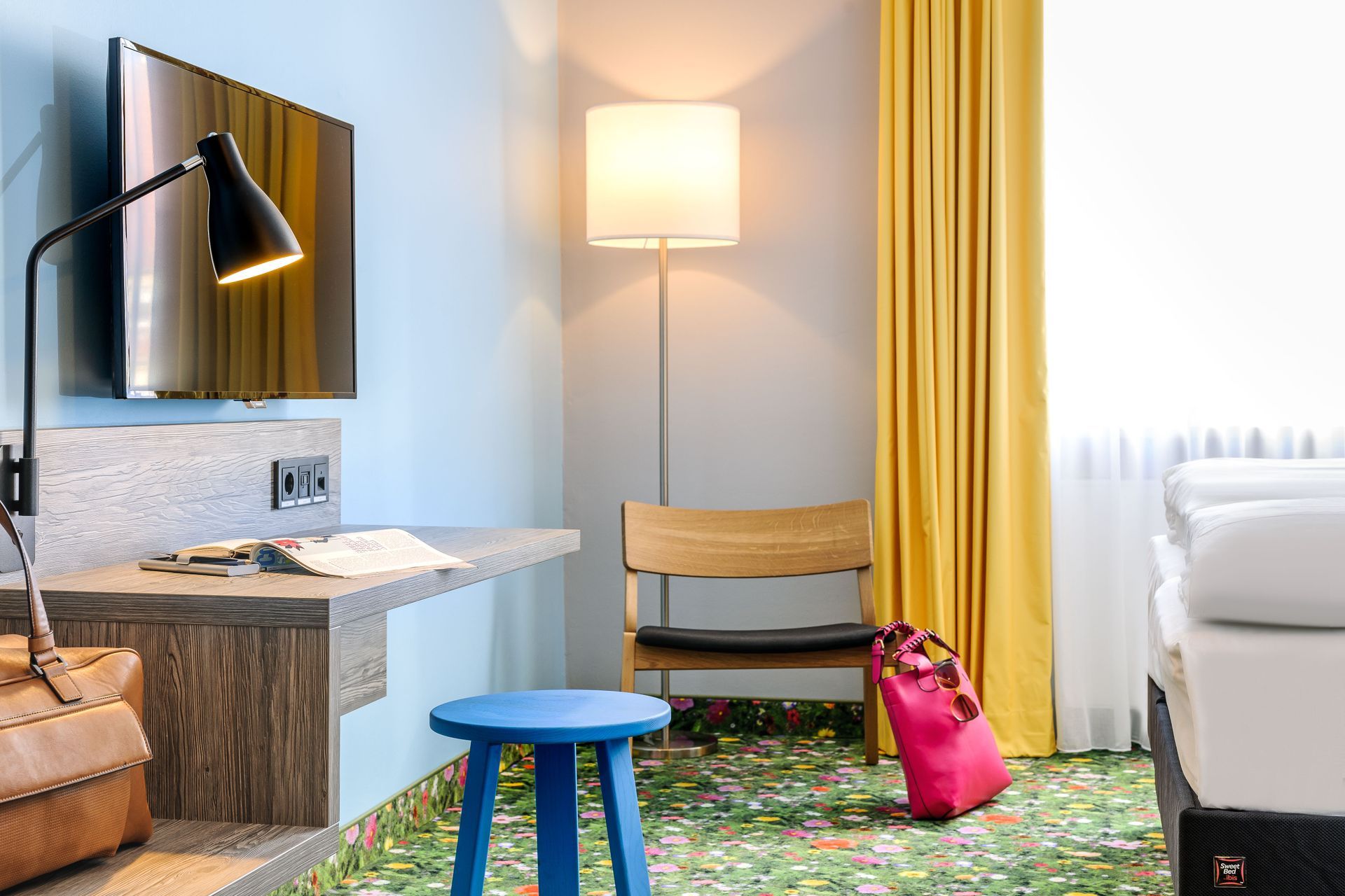 Hotelzimmer mit weißem Bett, Stehlampe, Schreibtisch, blauem Hocker, gelben Vorhängen und rosa Tasche auf dem Teppich