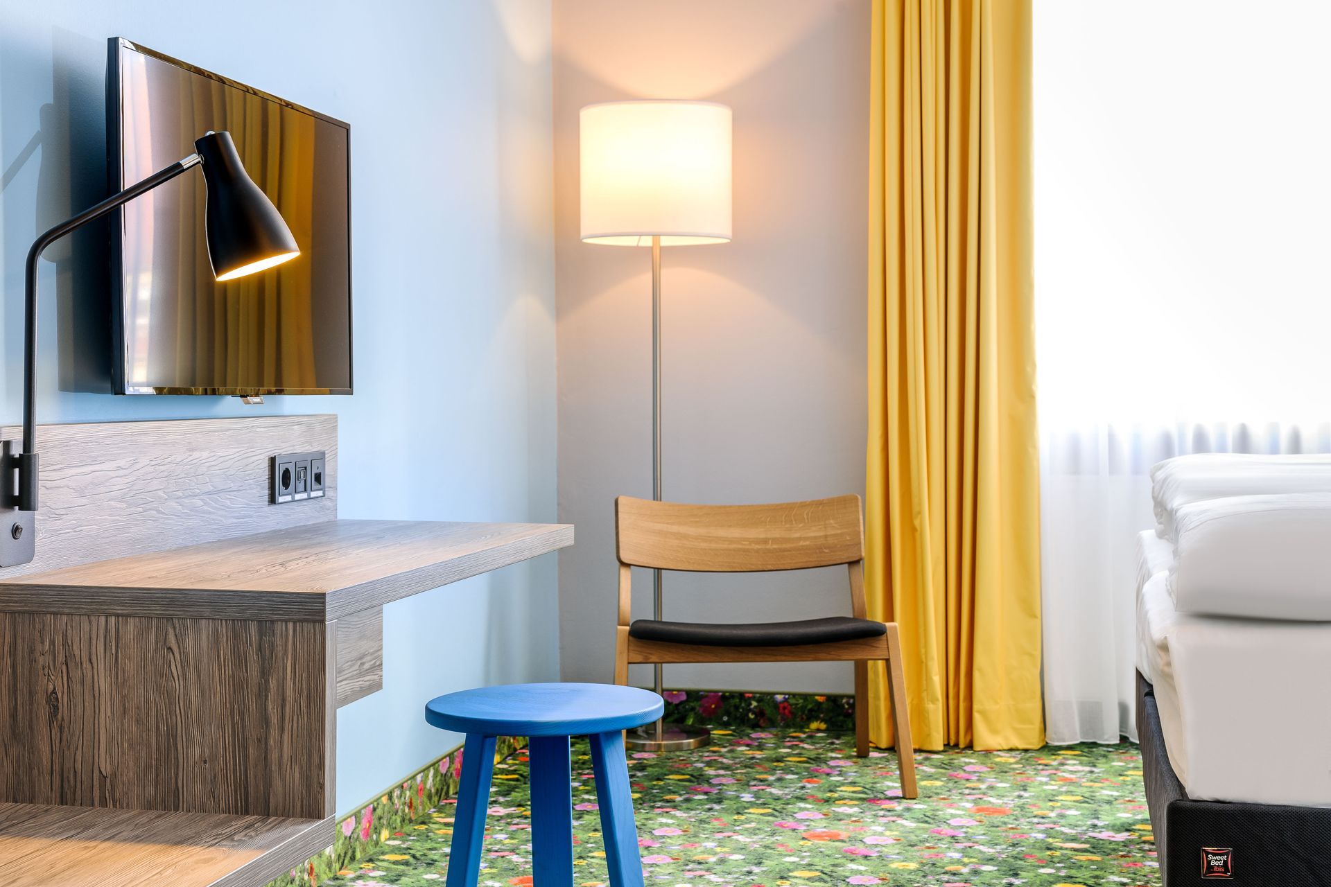 Modernes Hotelzimmer mit gelben Vorhängen, Stehlampe, Bett, Schreibtisch, blauem Hocker und gemustertem Teppich.