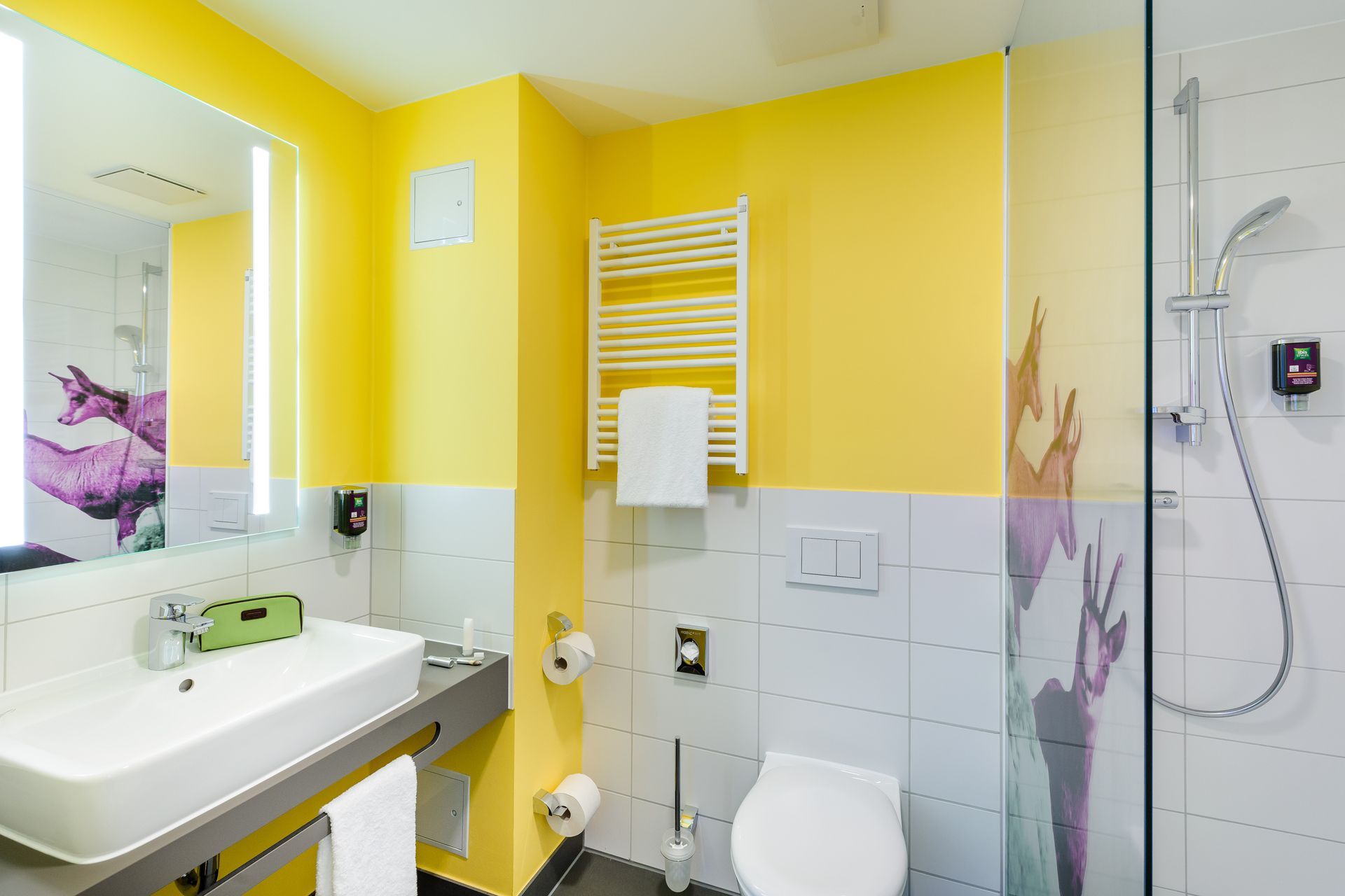 Hellgelbes Badezimmer mit Waschbecken, Toilette, Handtuchhalter und gefliestem Duschbereich mit wandmontiertem Duschkopf