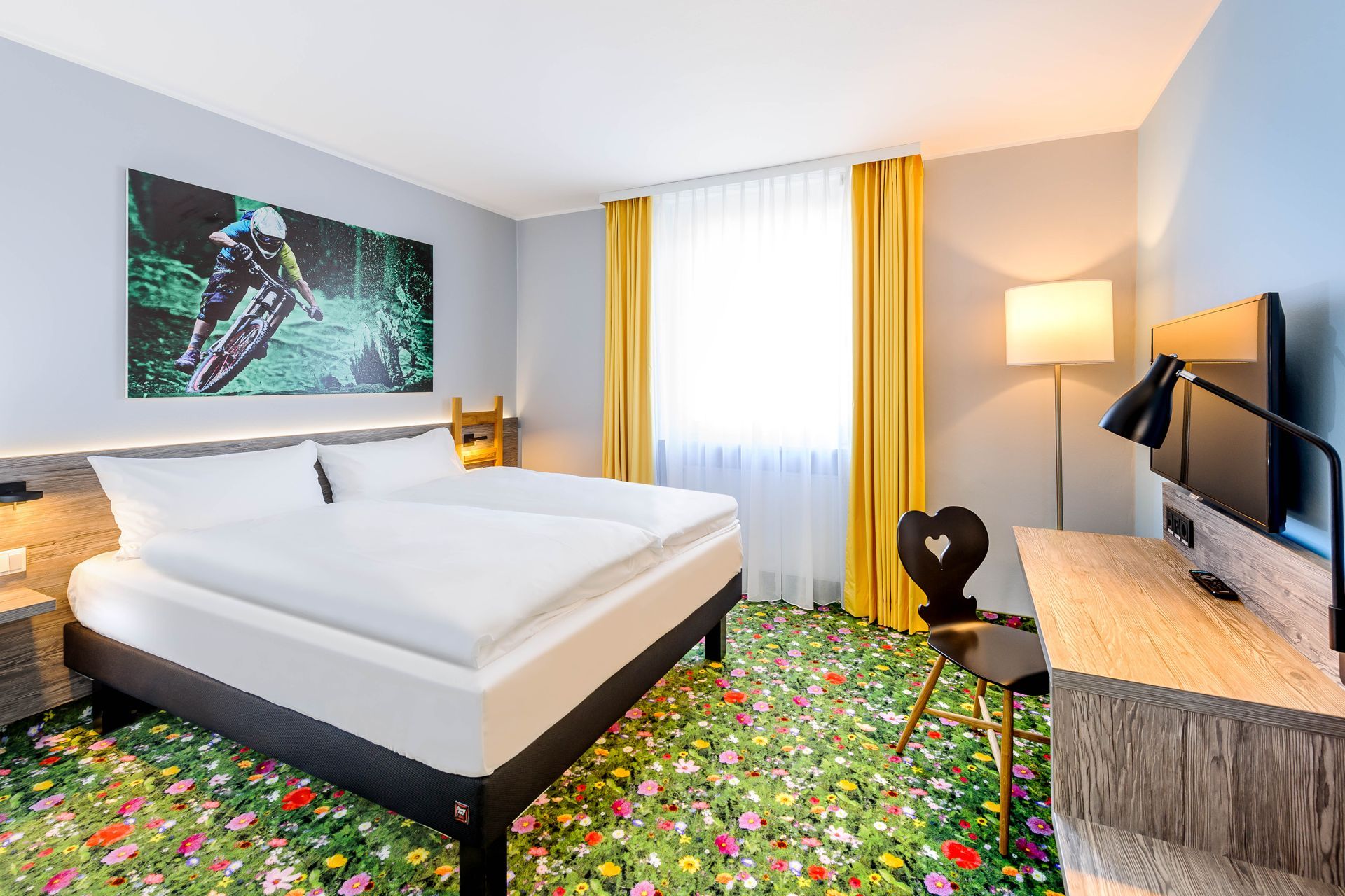 Modernes Hotelzimmer mit weißem Bett, buntem Blumenteppich, Fernseher und Fenster mit gelben Vorhängen.