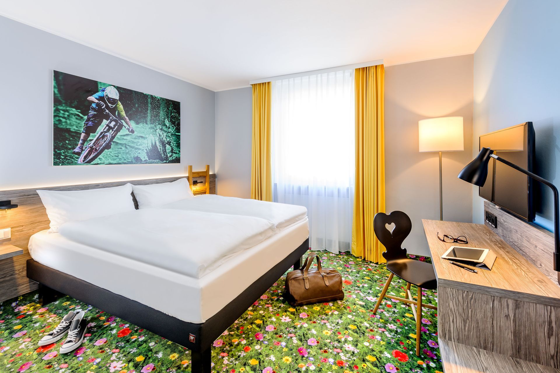 Modernes Hotelzimmer mit großem Bett, Wandbildern, Schreibtisch, Fernseher und farbenfrohem Blumenteppich.