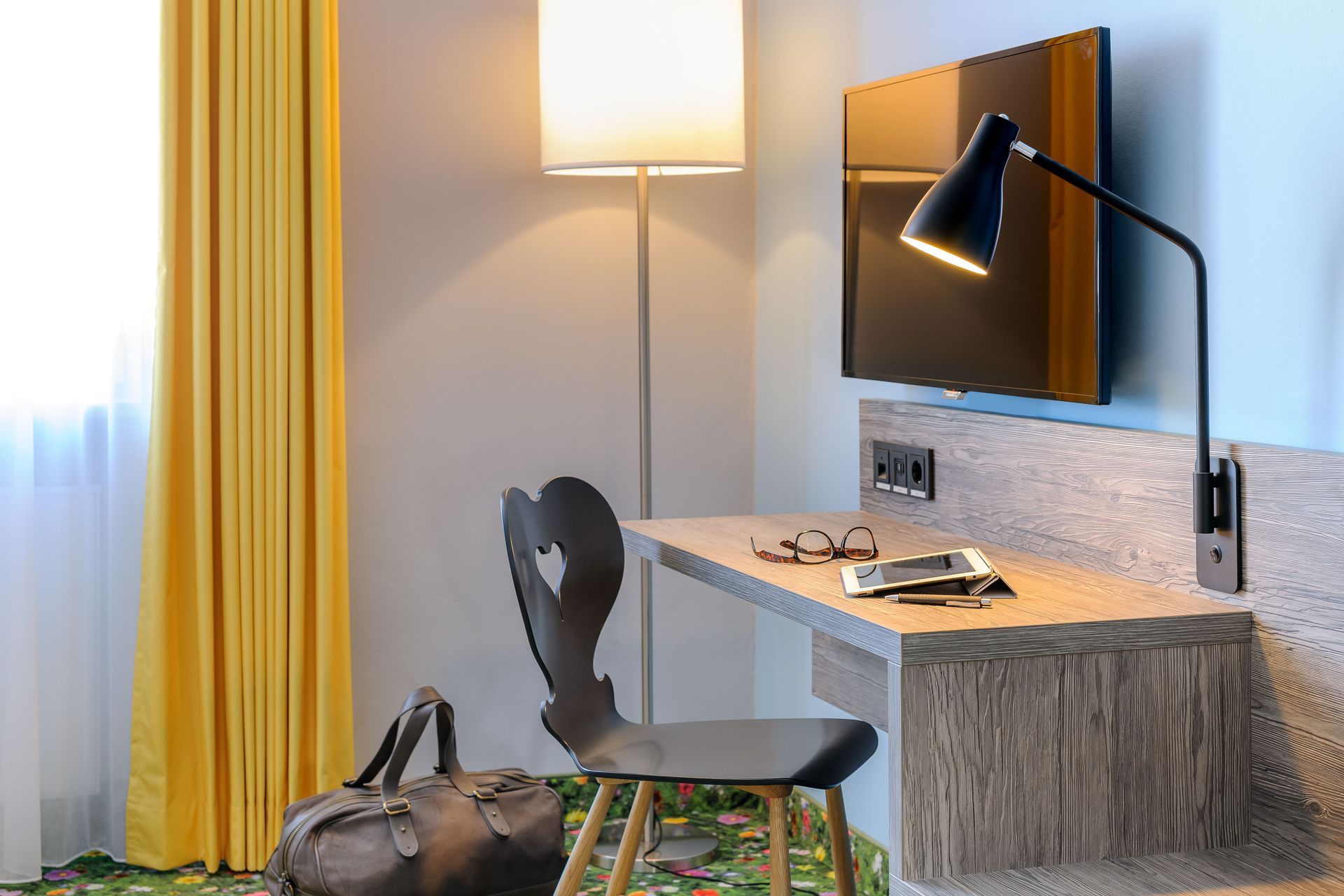 Moderner Schreibtisch mit Lampe, Wandfernseher, Stuhl und Taschen vor gelben Vorhängen.