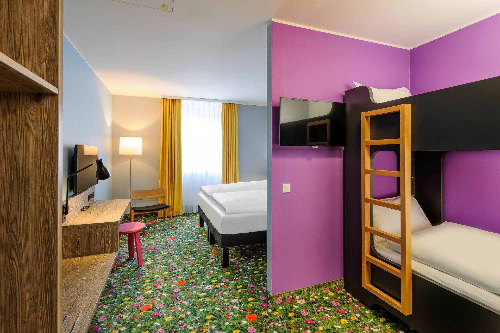 Kleines Hotelzimmer mit Etagenbetten, einem Einzelbett, Schreibtisch und lila Akzentwand, hell erleuchtet durch ein Fenster.