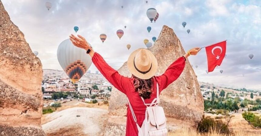 A person standing with arms outstretched, holding a Turkish flag, with hot air balloons floating in the sky in a scenic landscape.