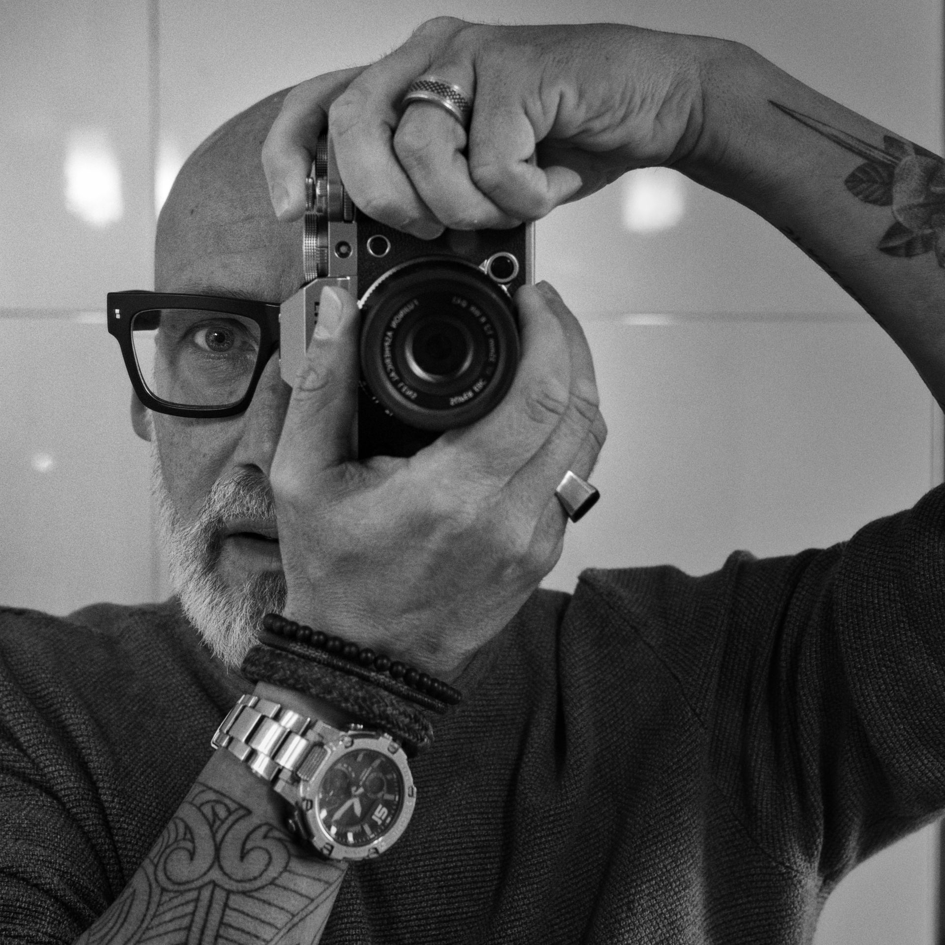 Man holding a camera to his face, taking a mirror selfie, with tattoos and a wristwatch visible.