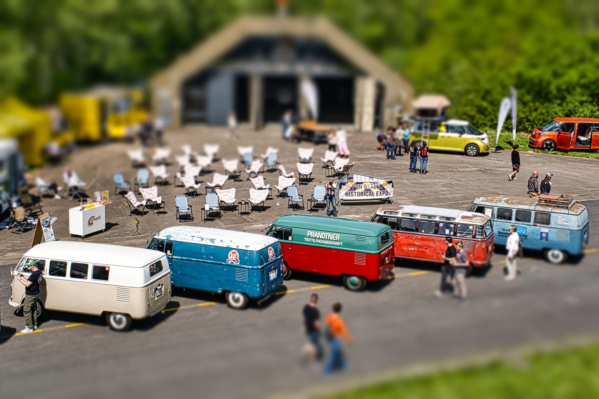 Miniature scene of colorful vans and people in front of a small building, with rows of tiny figures behind them