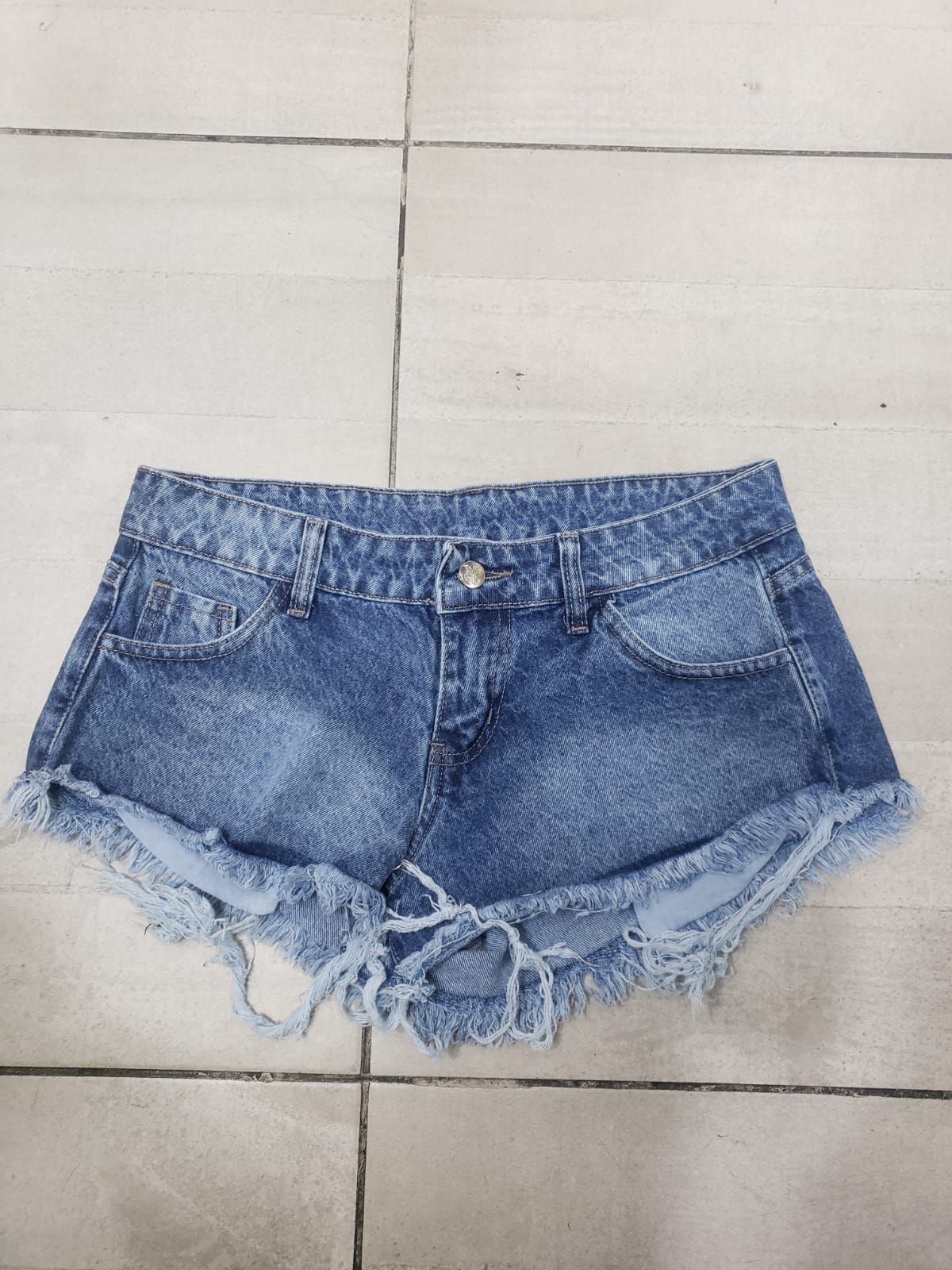 Pantalones cortos de mezclilla azul con bordes deshilachados, colocados sobre una superficie de baldosas.