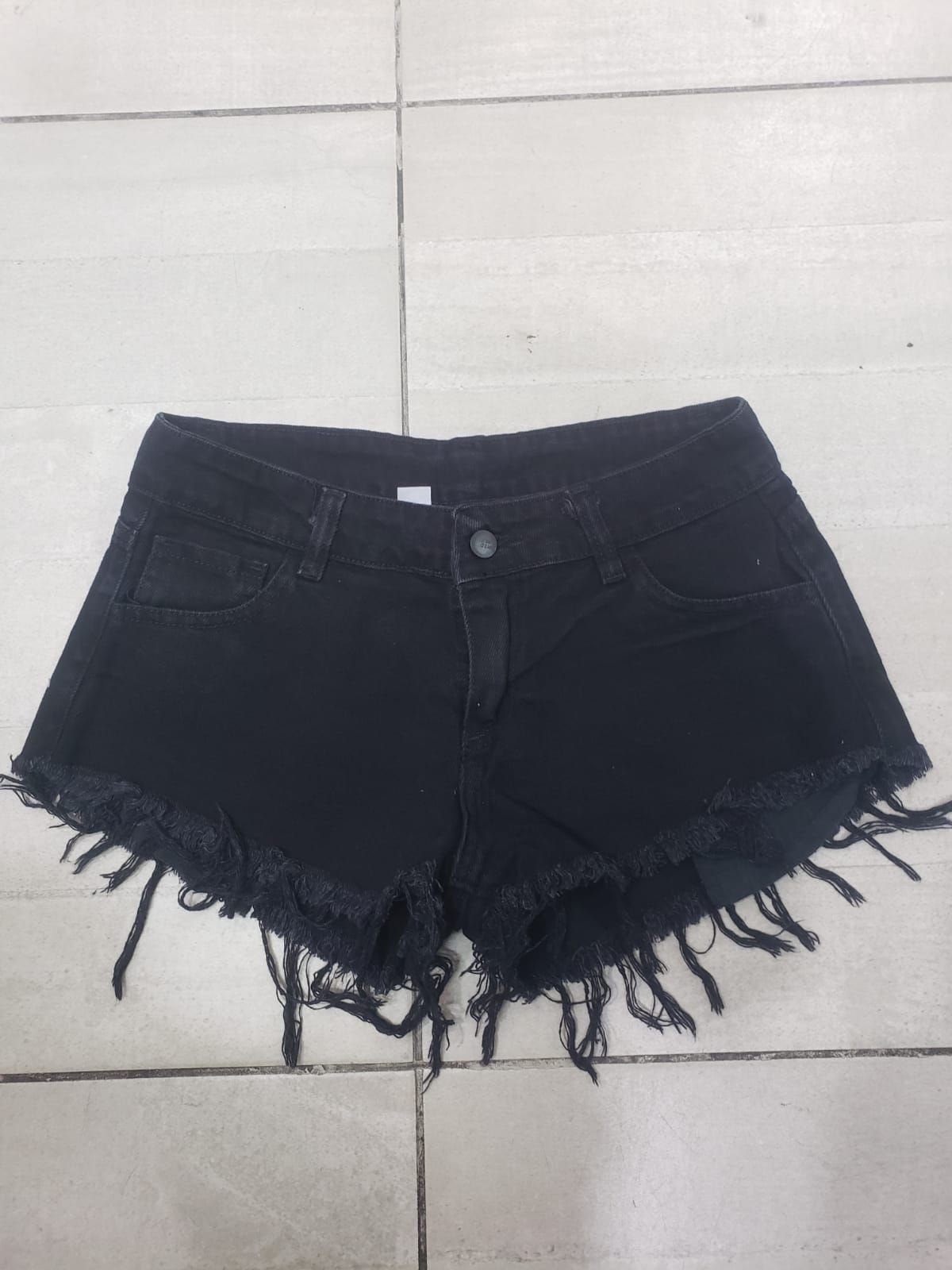 Pantalones cortos de mezclilla negros con bordes deshilachados, sobre un piso de baldosas.