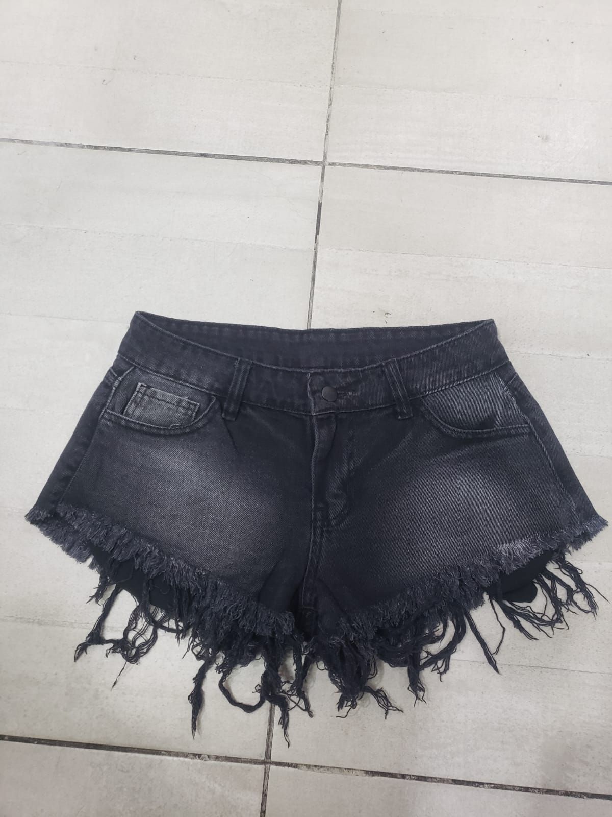 Pantalones cortos de mezclilla negros con bordes deshilachados sobre una superficie de baldosas.