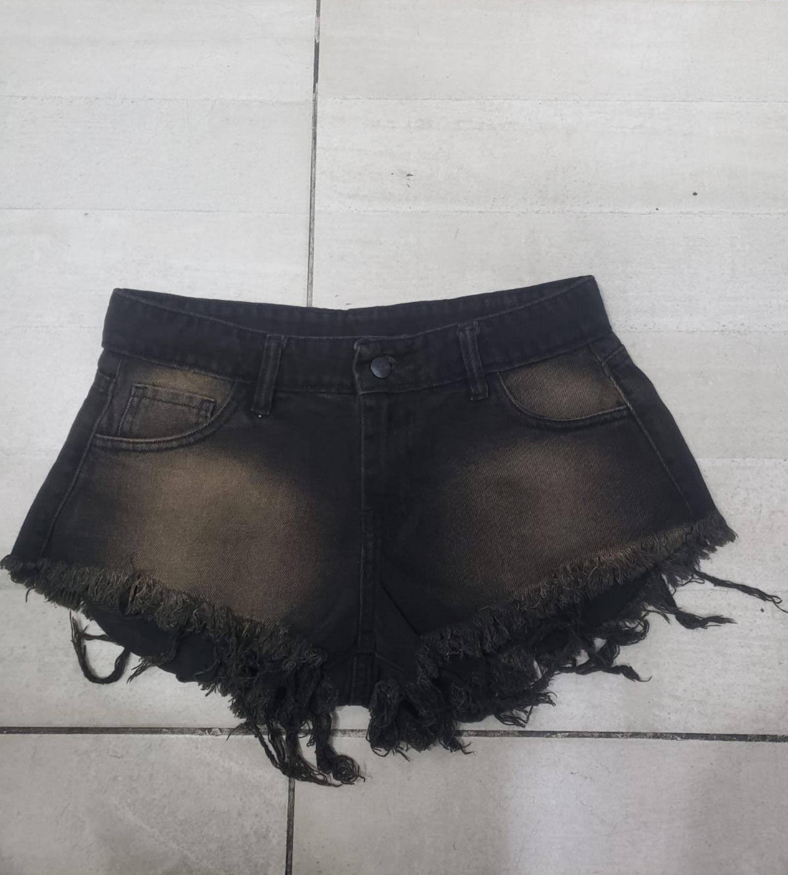 Shorts de mezclilla negros con bordes deshilachados y lavado desgastado sobre una superficie de baldosas.