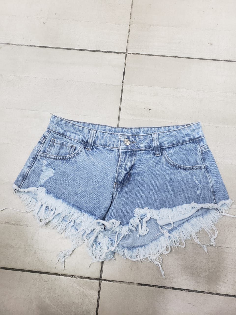 Pantalones cortos de mezclilla de lavado claro con bordes deshilachados y detalles desgastados sobre un piso de baldosas.
