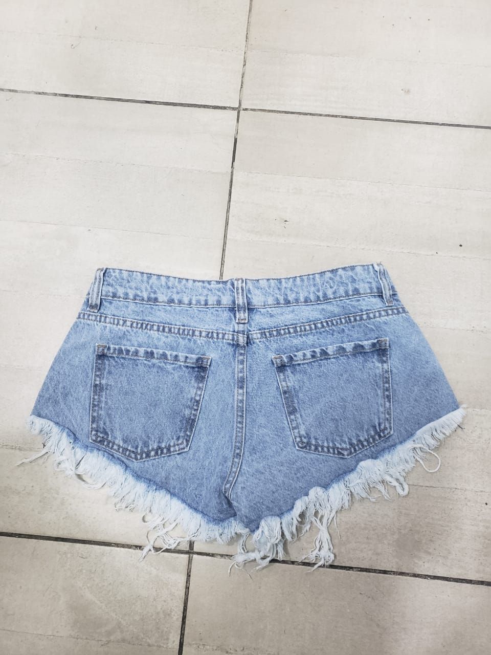 Pantalones cortos de mezclilla de lavado claro con bordes deshilachados y detalles desgastados sobre un piso de baldosas.