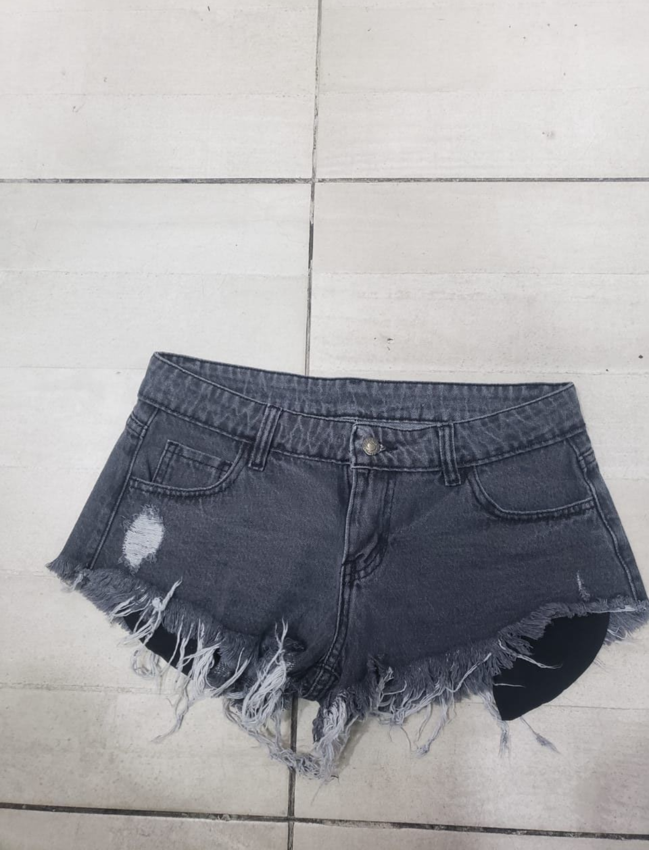 Pantalones cortos de mezclilla negros con bordes deshilachados y un parche desgastado sobre un piso de baldosas.