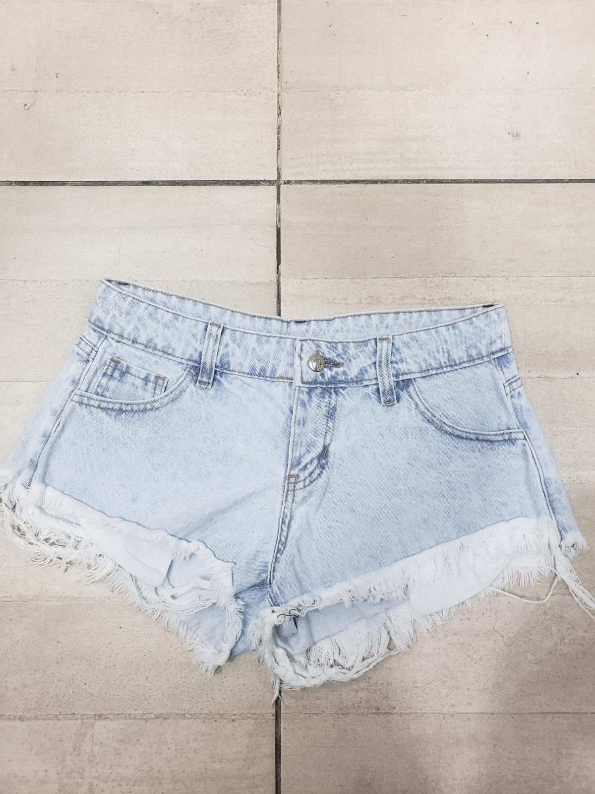 Pantalones cortos de mezclilla de lavado claro con bordes deshilachados, colocados sobre una superficie de baldosas.