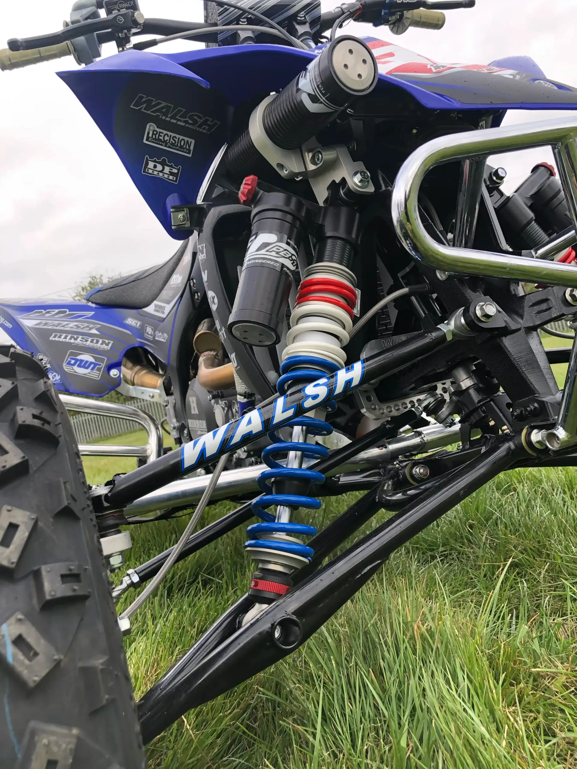 Blue and black ATV front suspension with a red, white, and blue shock absorber, outdoors on grass.