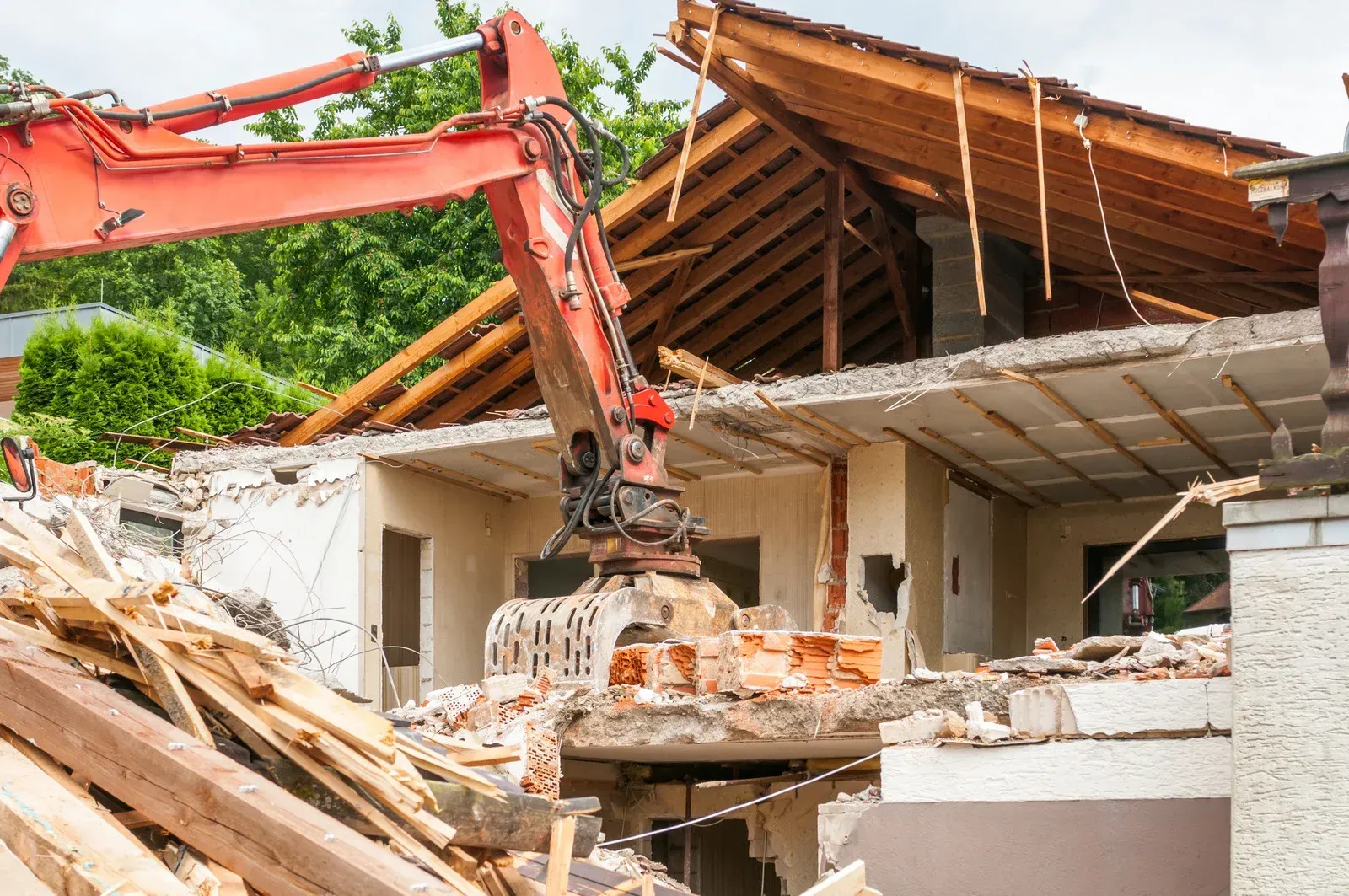 An excavator is demolishing a house with a pile of wood in the foreground.