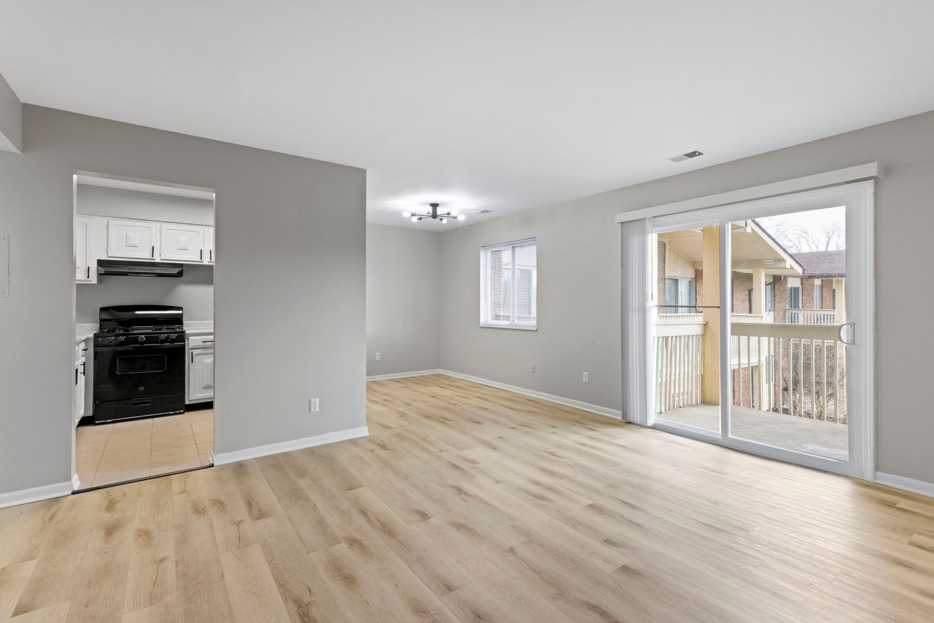 Empty apartment living room with light wood floors, sliding door to balcony, and view into the kitchen.