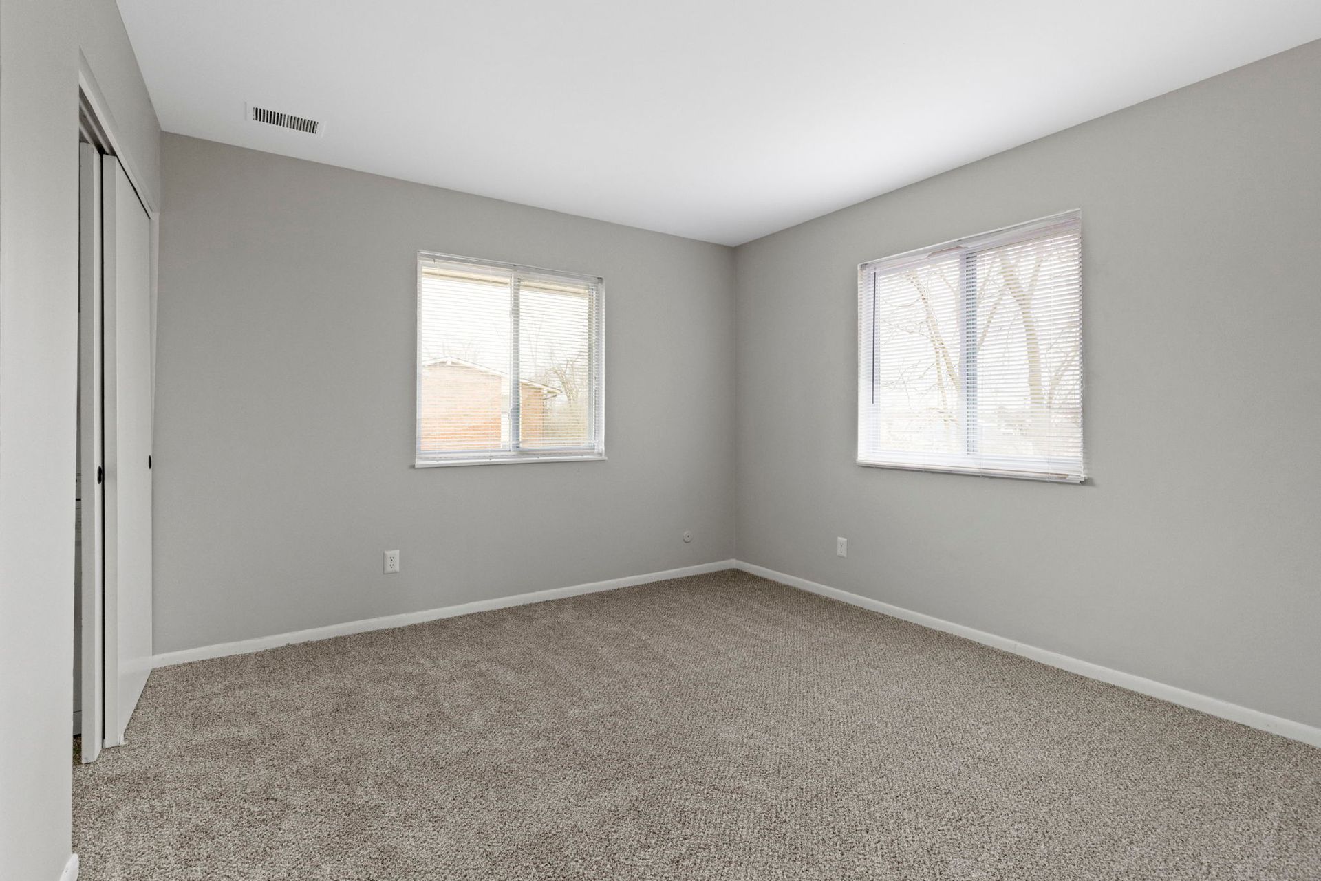 Empty bedroom with gray walls, carpet, two windows with blinds, and a closet.