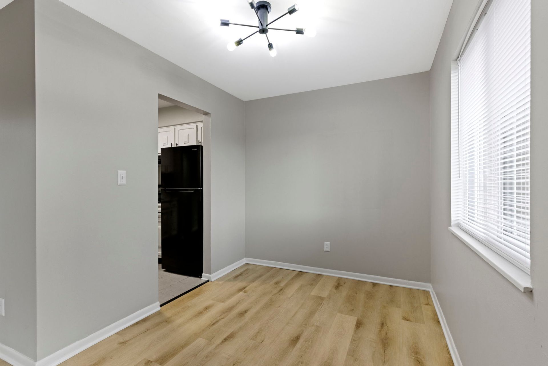 Empty dining room with gray walls, wood floors, and a window with blinds. A doorway leads to the kitchen.
