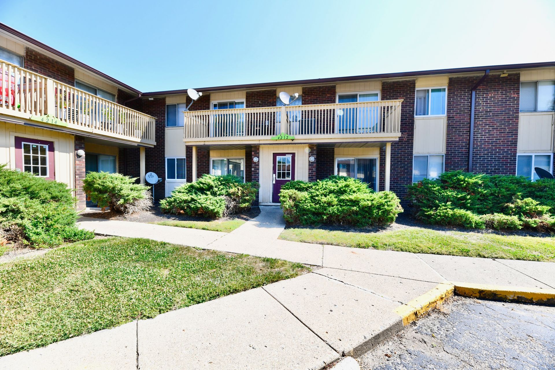 Apartment building exterior with brick and beige facade, walkways, and shrubs. Bright sunny day.