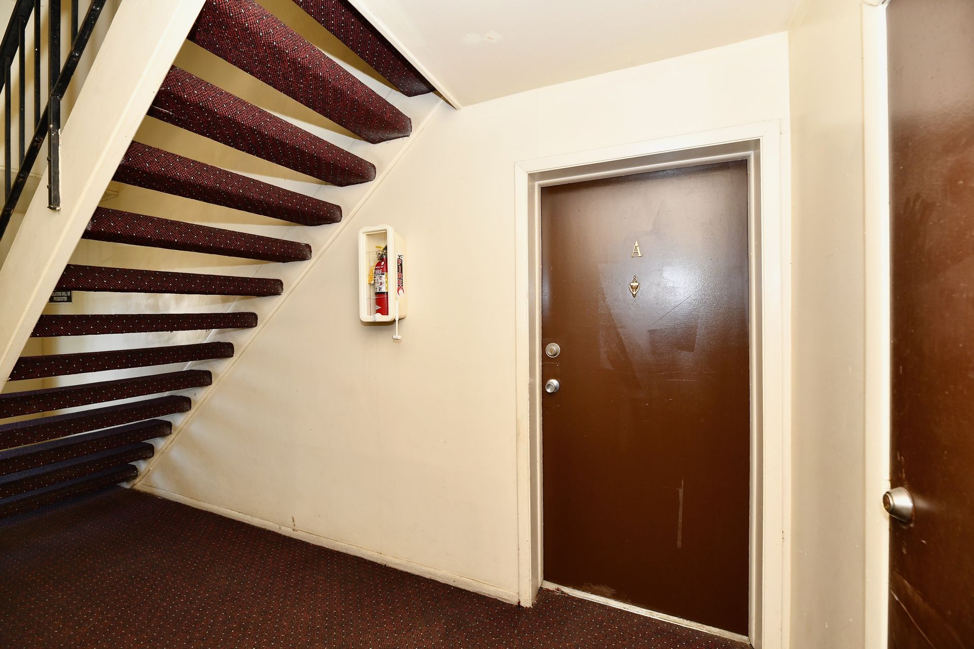 Stairwell with maroon carpet, brown door, fire extinguisher, and cream-colored walls.
