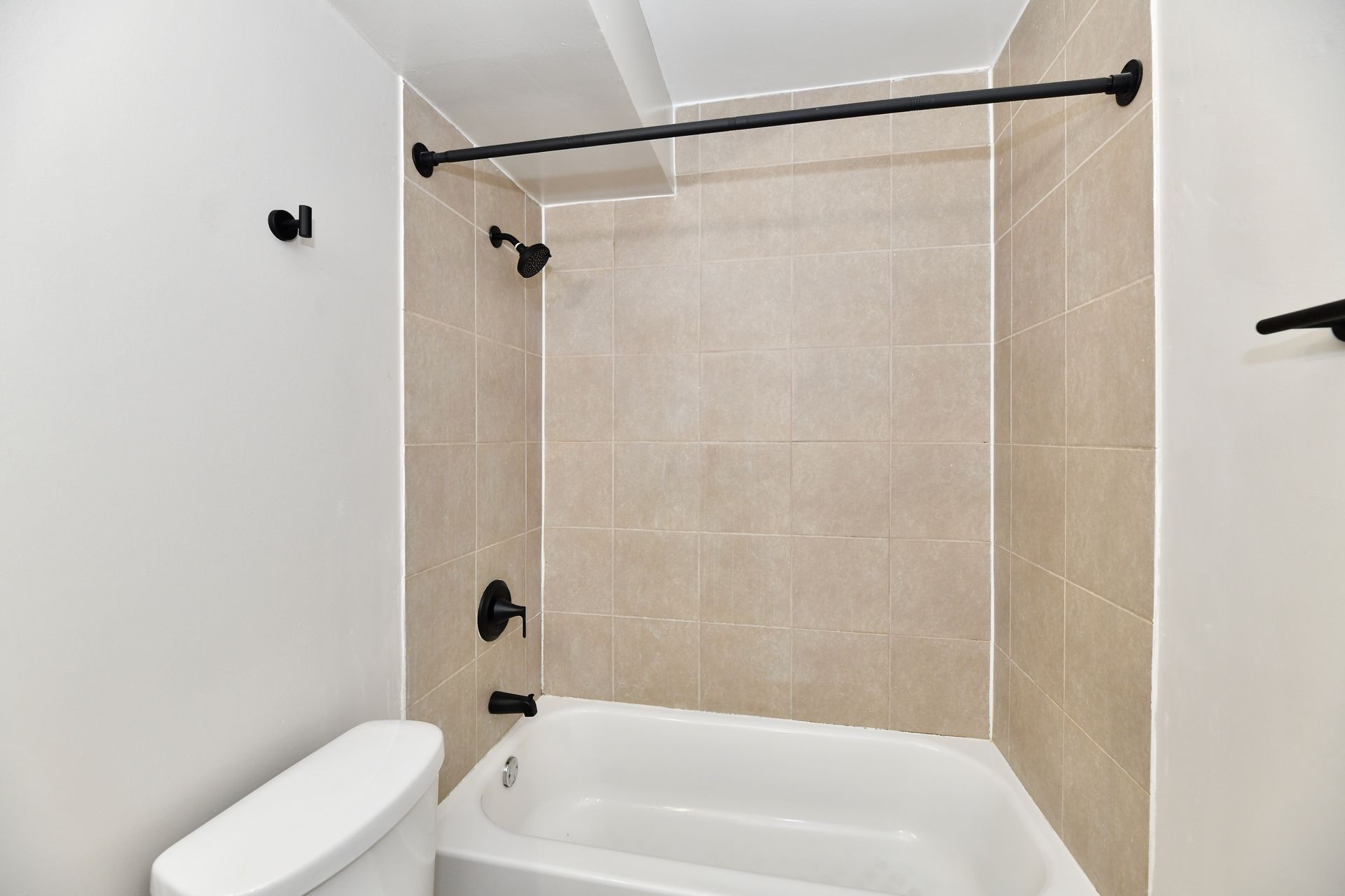 Bathroom with beige tiled shower, white tub and toilet, black fixtures, and shower rod.