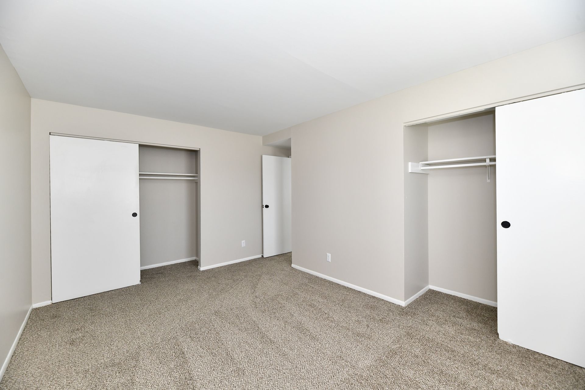Bedroom with neutral-colored walls, carpet, two closets with sliding doors, and a doorway in the background.
