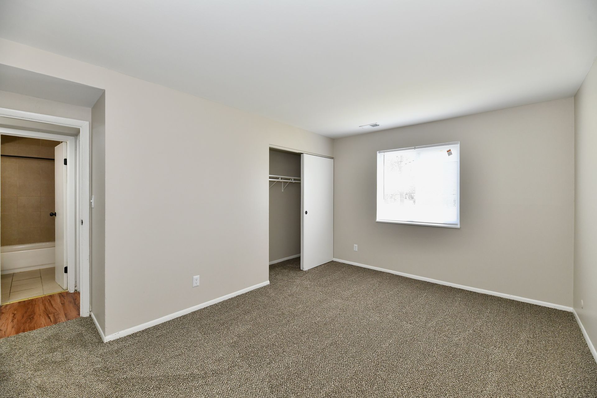 Bedroom with neutral-toned walls, brown carpet, white closet doors, and a small window.