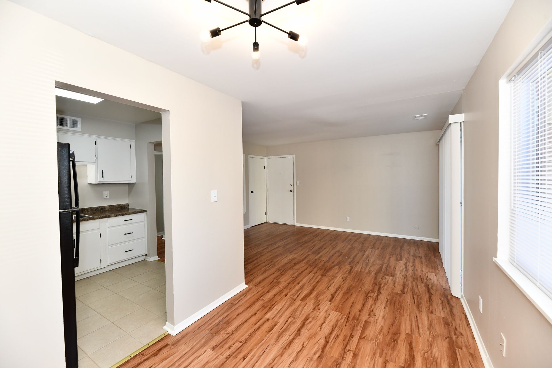 Empty living room with wood-look flooring, light walls, and an open kitchen doorway.