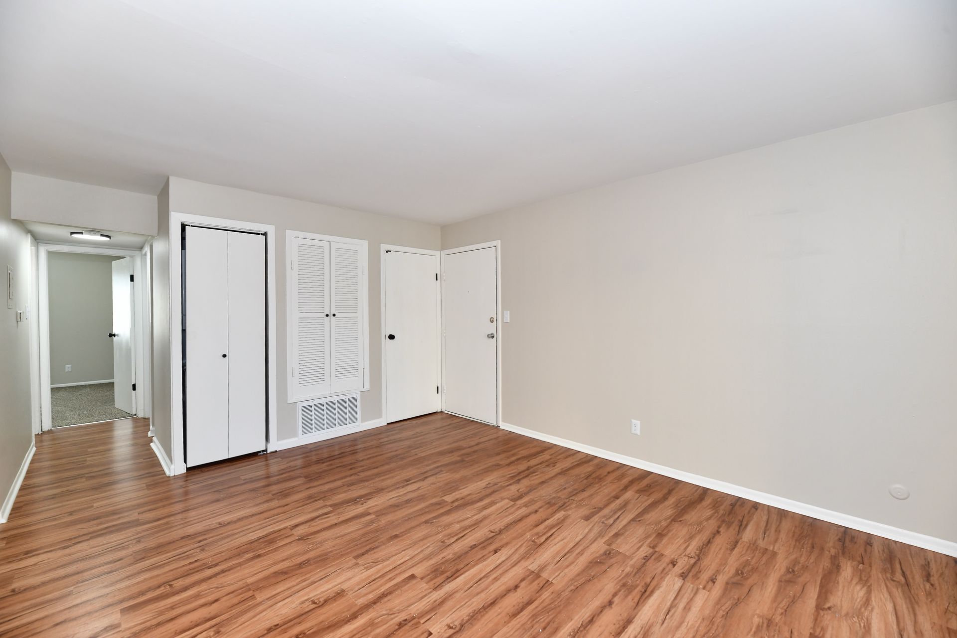 Empty room with wood-look flooring, white doors, and a hallway leading to another room.