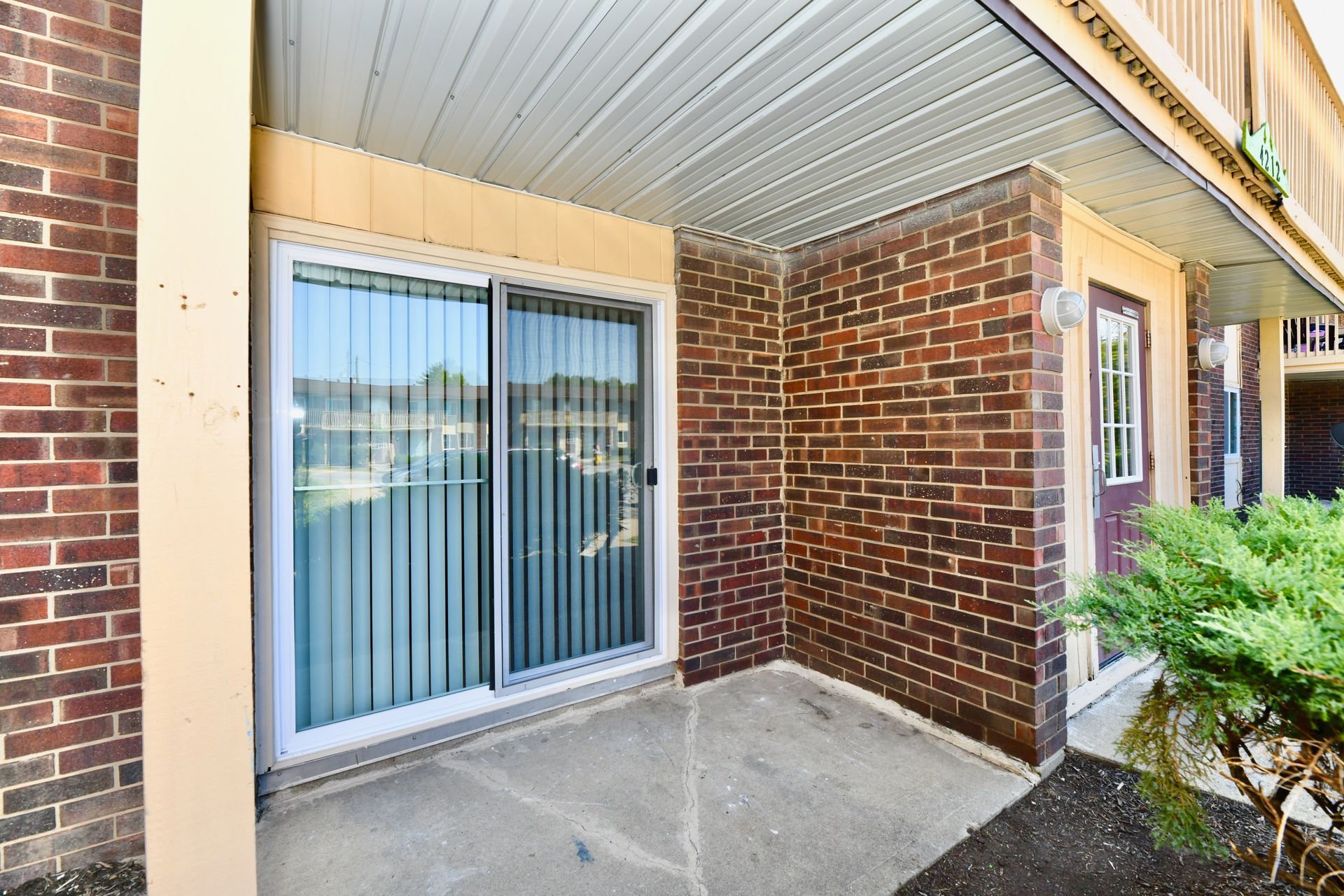 Covered patio with sliding glass door, brick walls, concrete floor, and a small bush.