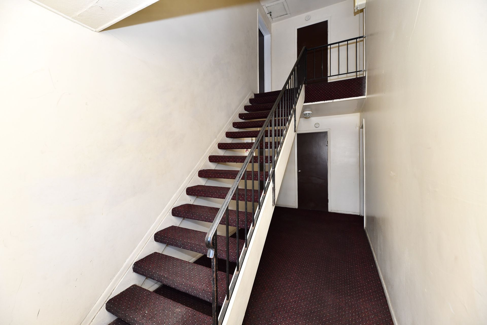 Stairwell with maroon carpeted stairs, black railing, and two closed doors on the landings. White walls.