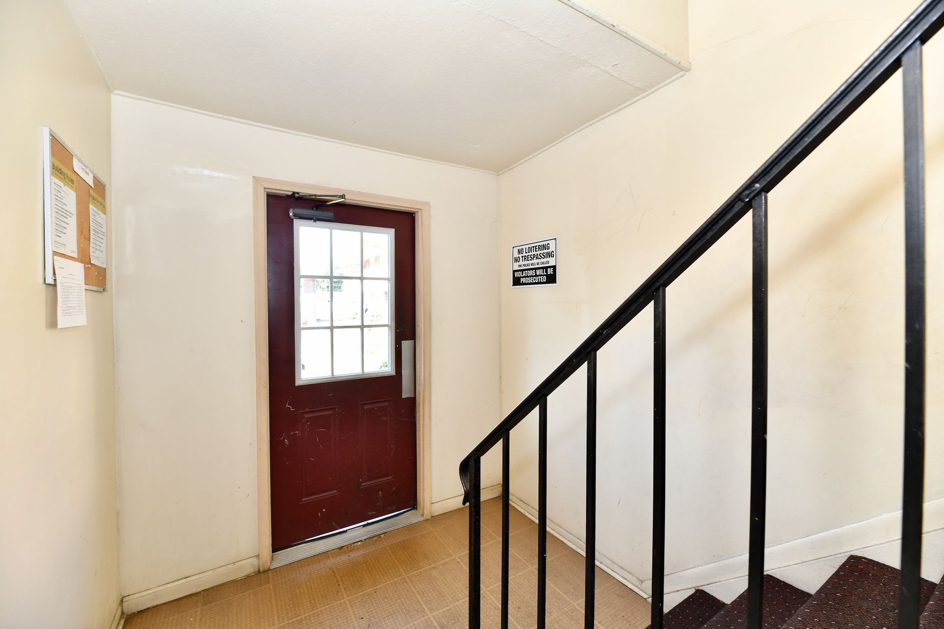 Dark red door at top of stairs with black railing, bulletin board on wall.