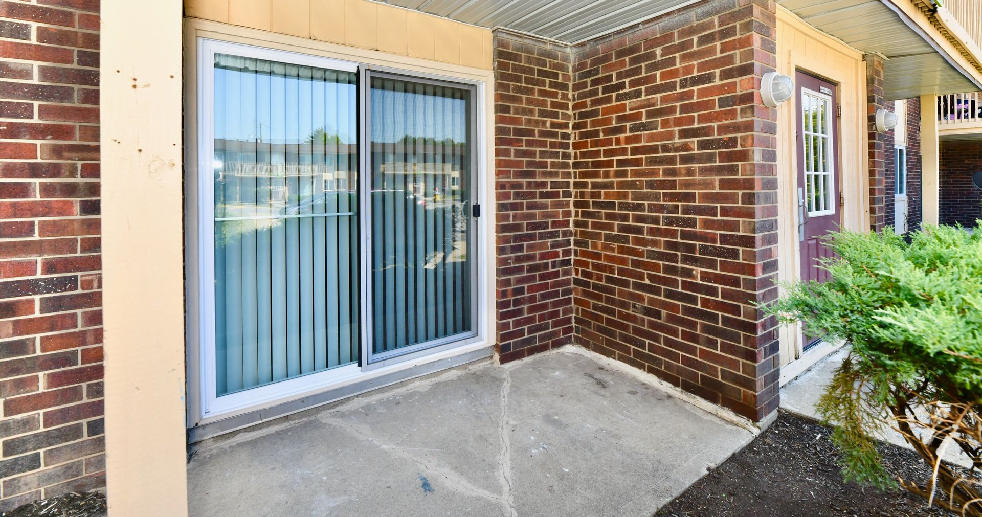 Exterior of a brick building with a sliding glass door and a small porch area, with greenery.