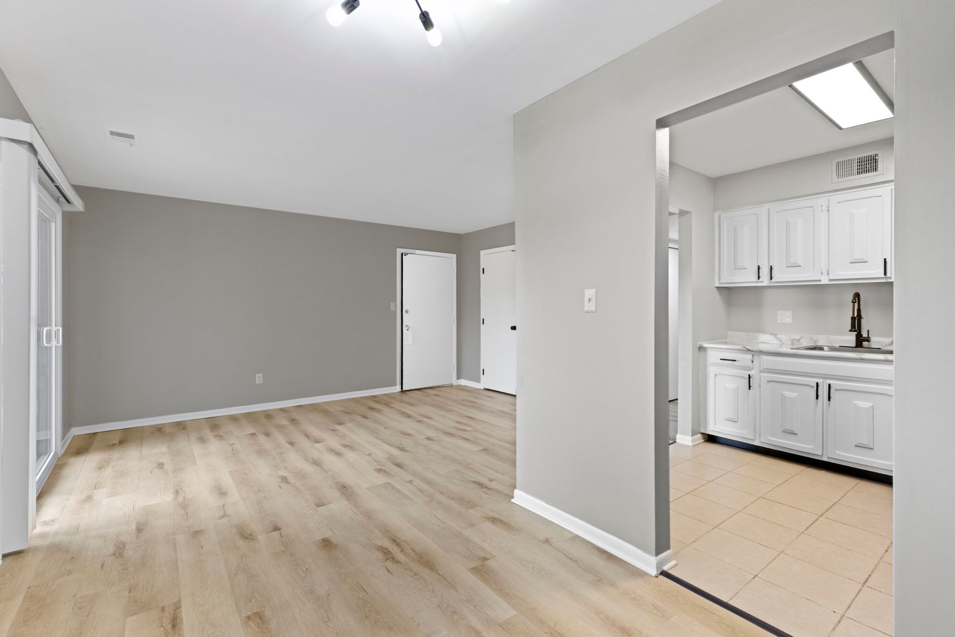 Empty living space with light wood floors, gray walls, and a view into a white-cabinet kitchen.