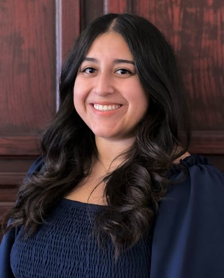 A woman in a blue shirt is smiling for the camera in front of a wooden wall.